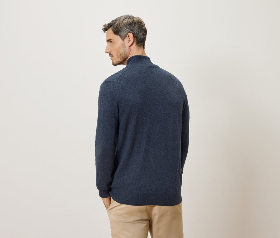 Un homme vu de dos portant un pull bleu marine et un pantalon beige sur un fond crème.