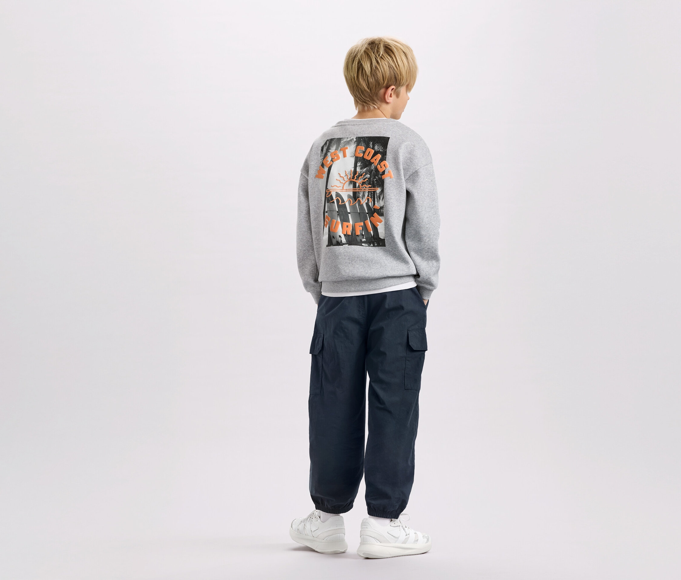 Un enfant de dos, portant un sweat-shirt gris avec l'inscription "West Coast Surfing", un pantalon cargo bleu marine et des baskets blanches.