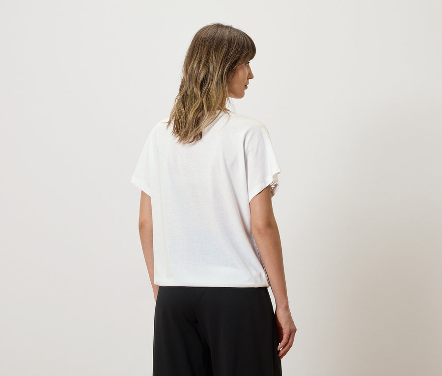 Une femme aux cheveux ondulés mi-longs porte un t-shirt blanc avec des applications en dentelle et un pantalon noir.