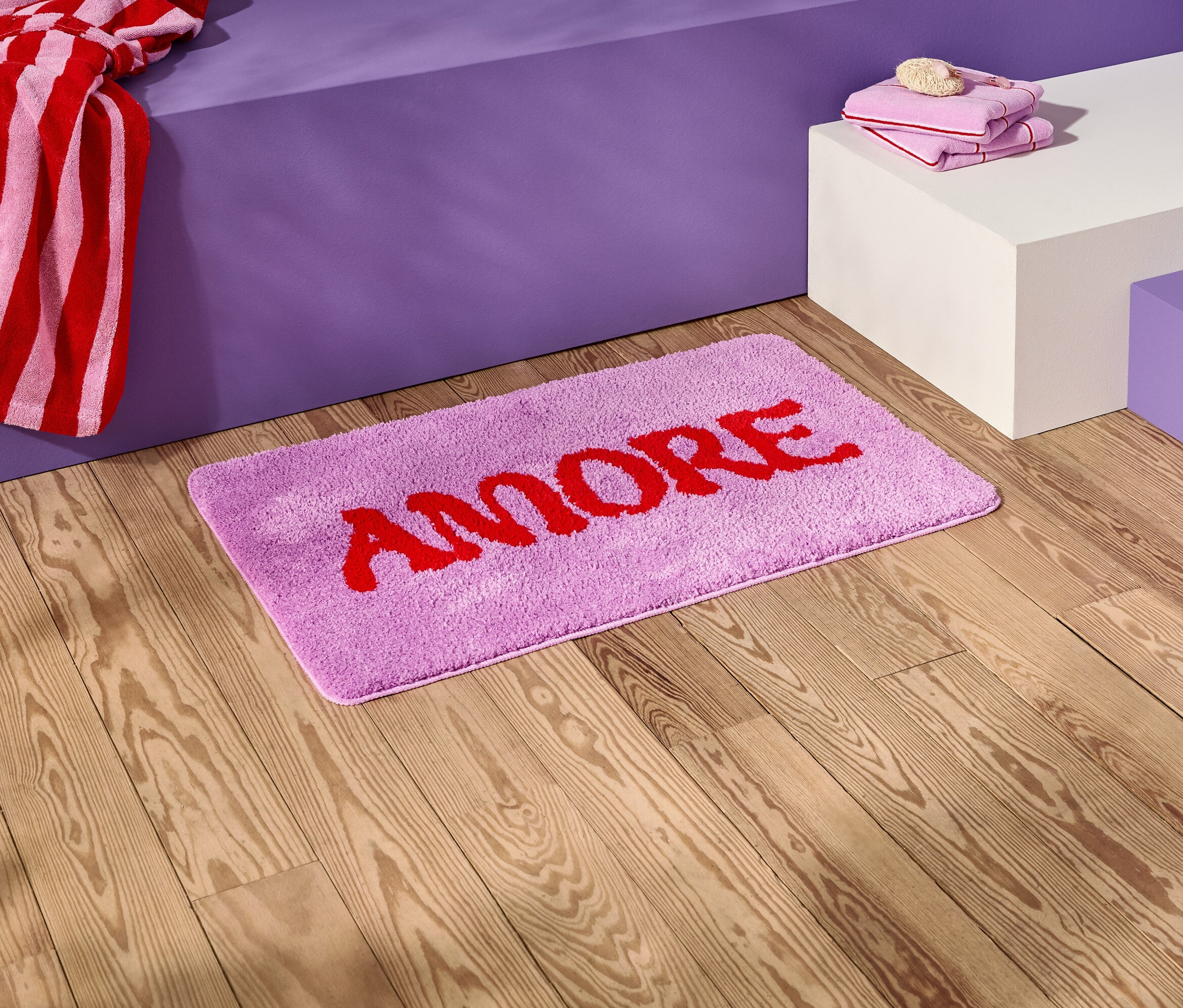 Sur un plancher en bois se trouve un tapis de bain lilas avec l'inscription rouge "AMORE".