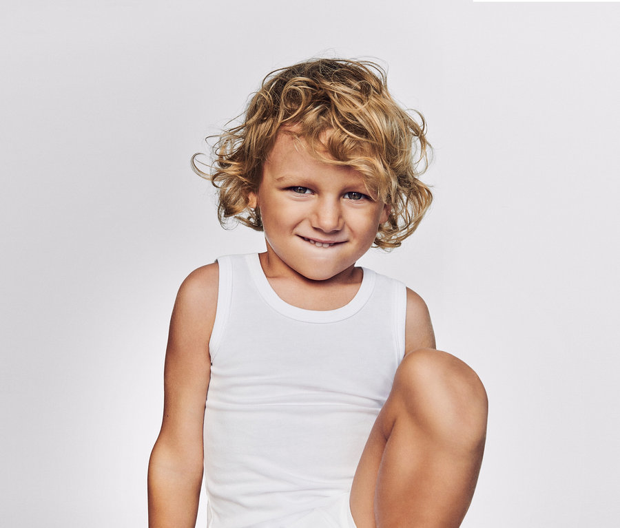 Enfant blond aux cheveux bouclés, souriant et portant un débardeur blanc.