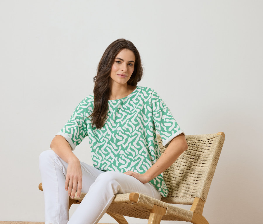 Femme assise sur une chaise, portant un haut vert à motifs blancs et un pantalon blanc.