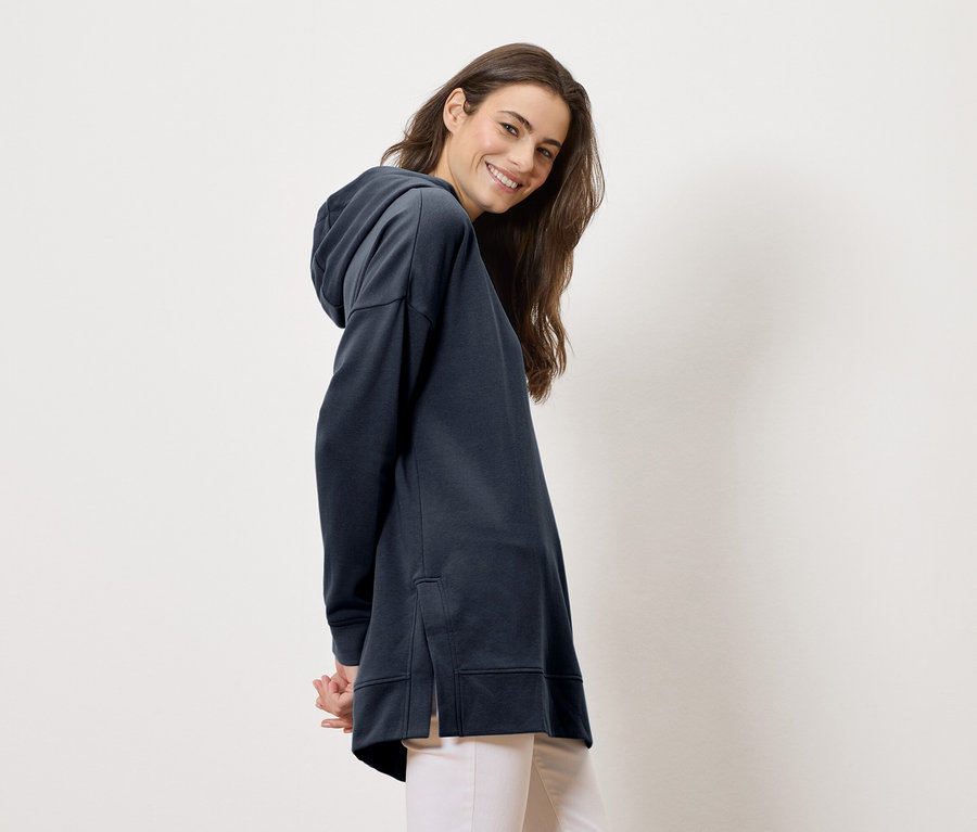 Femme portant un long sweat à capuche bleu marine et un pantalon clair, souriant à la caméra.