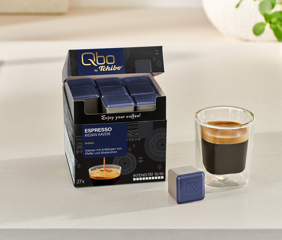 Un paquet ouvert de capsules Qbo Espresso INDIAN KAVERI, une capsule individuelle et un verre d'espresso sont posés sur une surface claire.