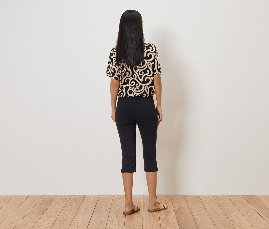Une femme aux cheveux foncés, vue de dos, porte un haut à motifs, un pantalon Capri noir et des sandales dorées devant un mur blanc.