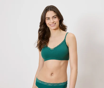Une femme souriante aux longs cheveux bruns portant un soutien-gorge vert avec de la dentelle et des sous-vêtements assortis sur fond blanc.