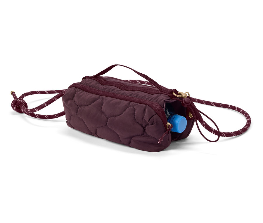 Un sac cylindrique matelassé aubergine avec cordon de serrage et une bouteille d'eau bleue qui dépasse de la fermeture éclair.