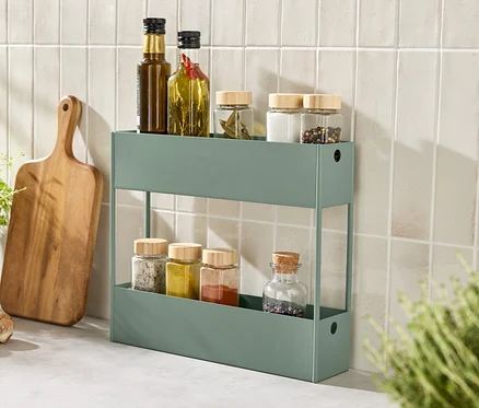 Une étagère verte avec des épices et des huiles est posée sur un comptoir de cuisine à côté d'une planche à découper et d'herbes aromatiques.