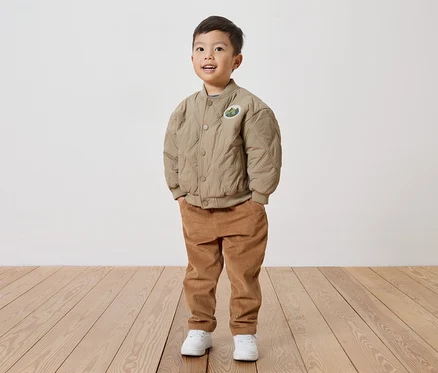 Un garçon souriant portant une veste matelassée beige avec un écusson, un pantalon en velours côtelé et des chaussures blanches se tient sur un plancher en bois devant un mur blanc.