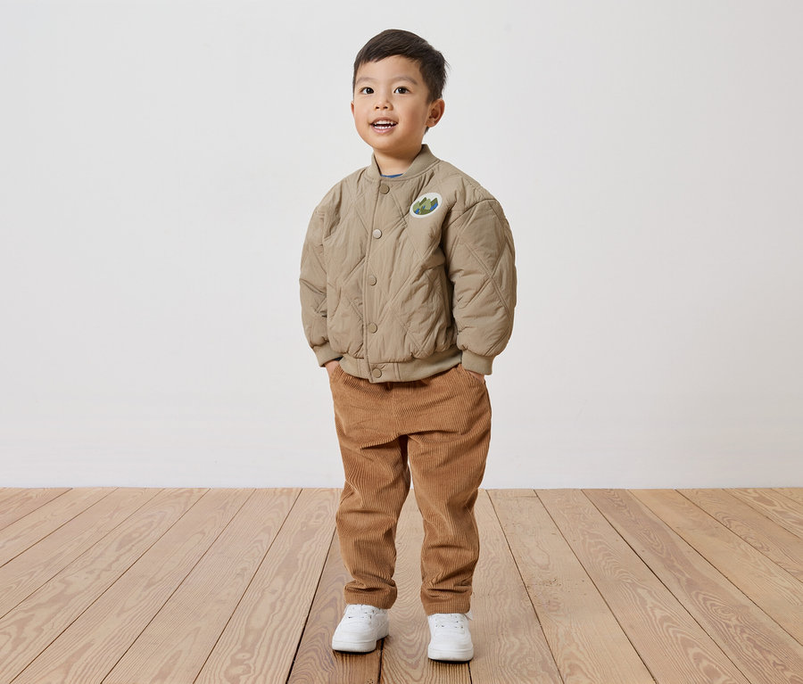Un garçon souriant portant une veste matelassée beige avec un écusson, un pantalon en velours côtelé et des chaussures blanches se tient sur un plancher en bois devant un mur blanc.