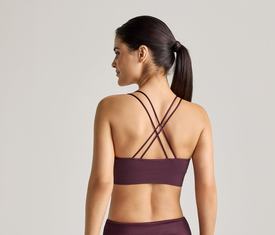 Femme vue de dos, portant un soutien-gorge de sport violet à bretelles croisées et un legging sur un fond clair.