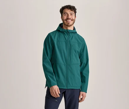 Un homme souriant aux cheveux bouclés et barbu se tient de face, portant une veste softshell verte à capuche et un pantalon bleu foncé.