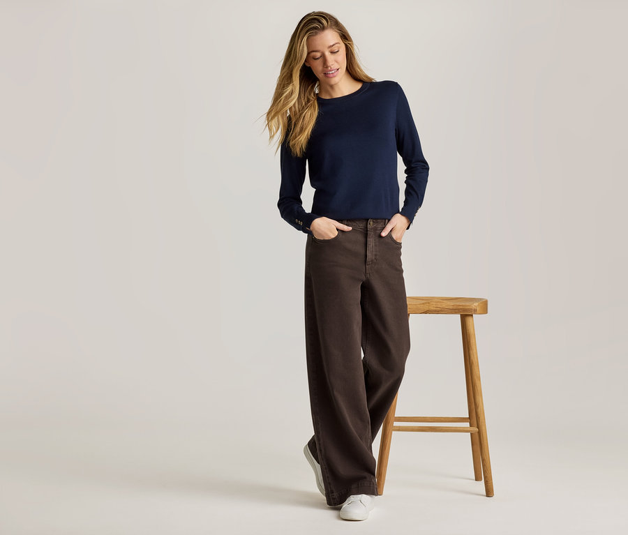 Une femme portant un pull bleu marine et un pantalon marron pose à côté d'un tabouret sur un fond clair.