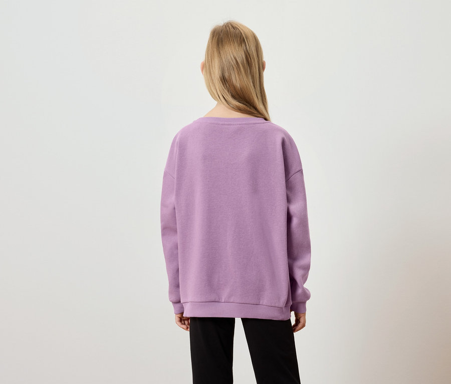 Vue arrière d'une jeune fille aux longs cheveux blonds, portant un pull lilas et un pantalon noir sur un fond neutre.