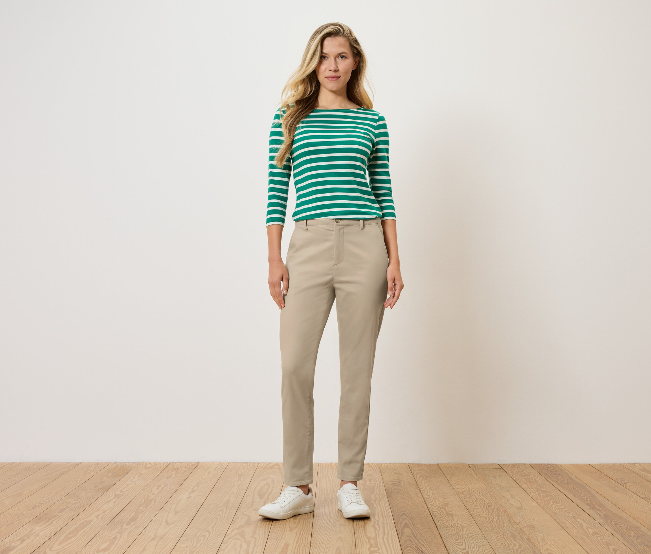 Une femme se tient debout et porte un haut à manches longues rayé vert et blanc, un pantalon beige et des chaussures blanches.