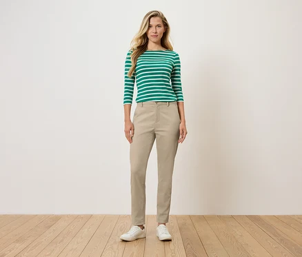 Une femme se tient debout et porte un haut à manches longues rayé vert et blanc, un pantalon beige et des chaussures blanches.