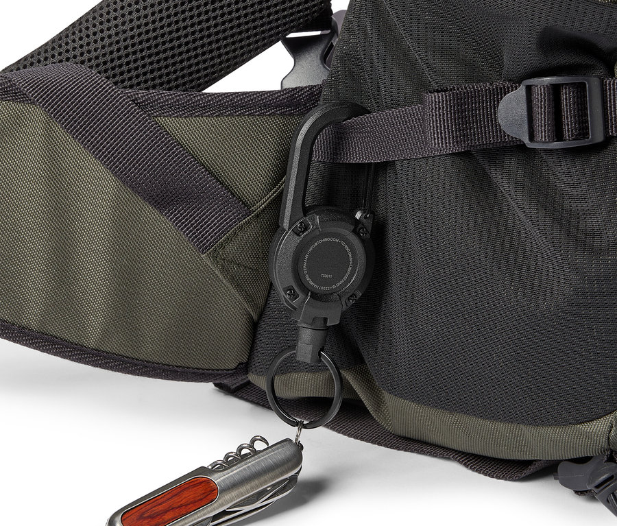Gros plan d'un sac à dos olive et noir avec un mousqueton noir auquel sont attachés un porte-clés et un couteau de poche argenté avec un manche en bois.