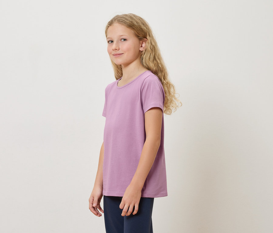 Une fille aux longs cheveux blonds portant un t-shirt violet et un pantalon bleu marine se tient devant un fond blanc.
