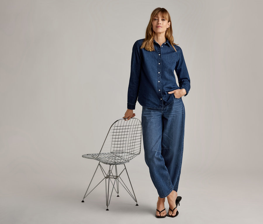 Femme en chemise et jean larges en denim, debout à côté d'une chaise en fil métallique, une main posée dessus et l'autre dans sa poche, portant des tongs à talons noires.