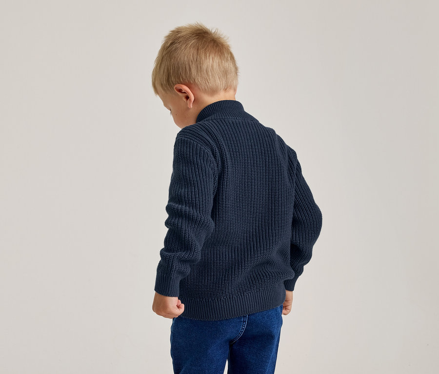 Jeune garçon de dos, portant un pull en maille bleu marine et un jean bleu, la tête légèrement baissée.