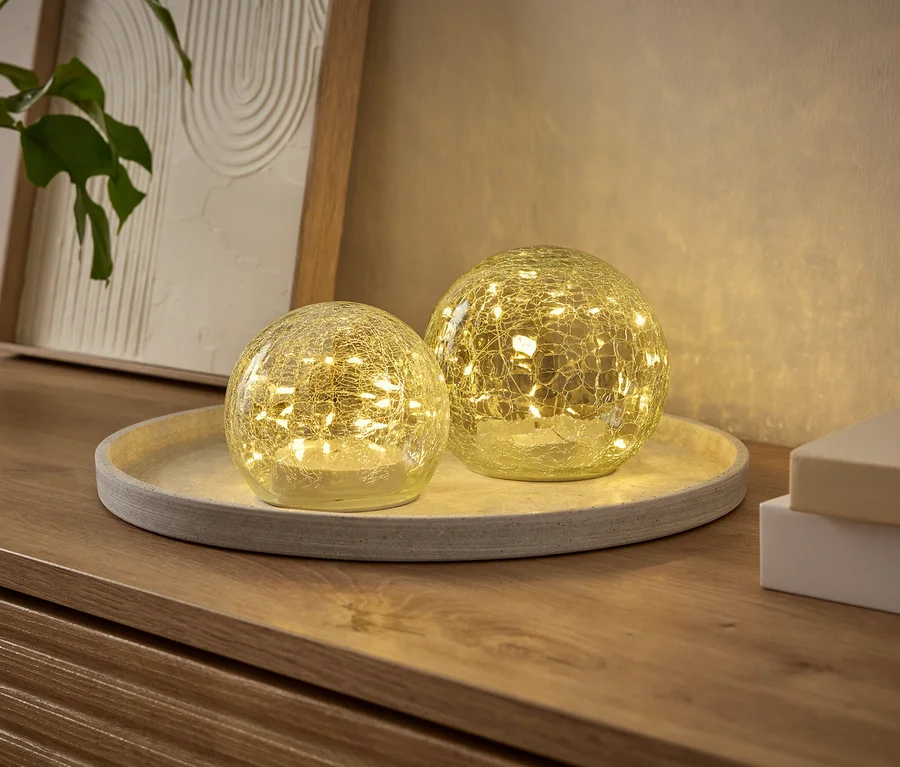 Deux lumières décoratives sphériques lumineuses avec un effet de verre craquelé, remplies de petites LED, sont posées sur un plateau rond sur une commode en bois. Une plante et un cadre sont en arrière-plan.
