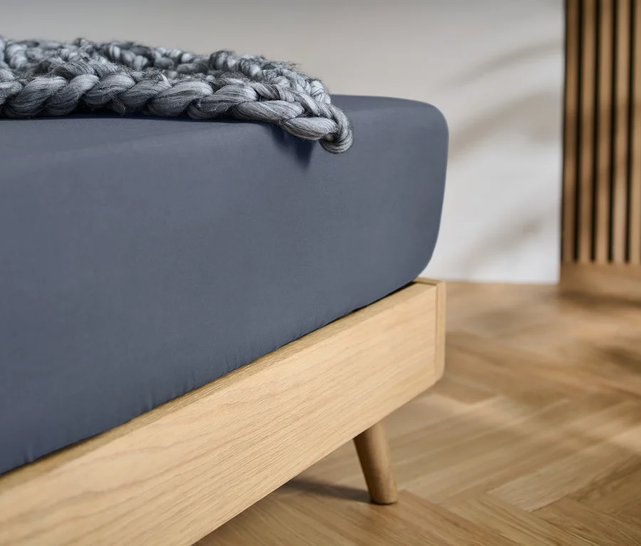 Un lit avec un cadre en bois clair, un drap-housse gris foncé et une couverture en tricot épais grise. Un mur à lattes en bois et un parquet clair sont en arrière-plan.