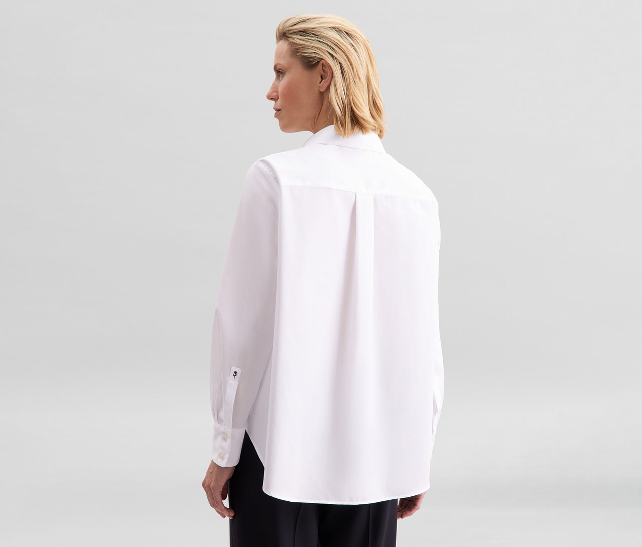 Femme vue de dos, la tête tournée vers la droite, portant un chemisier sans repassage Seidensticker blanc avec un logo noir sur le poignet gauche et un pantalon noir.