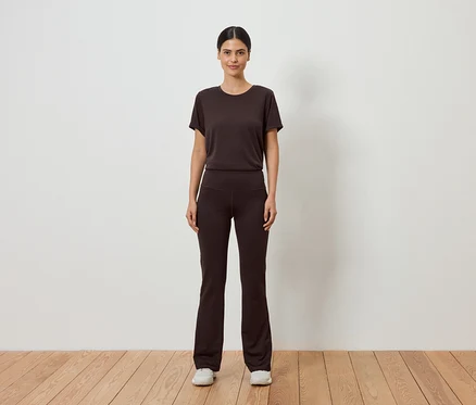 Une femme en t-shirt marron et pantalon évasé se tient devant un mur blanc sur un plancher en bois.