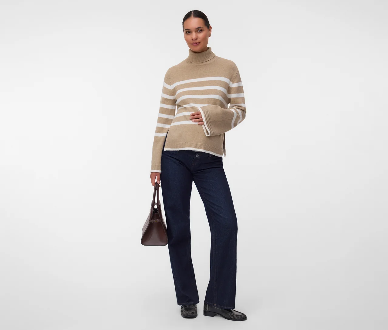 Femme debout, portant un pull col roulé beige à rayures blanches, un jean large bleu foncé et des mocassins noirs, tenant un sac à main marron.