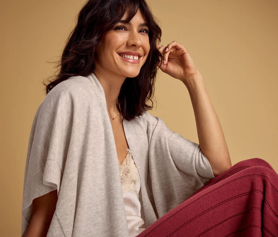 Femme souriante aux cheveux foncés et frange, portant un cardigan ouvert gris clair sur un débardeur en dentelle crème et un pantalon rayé rouge foncé, regardant vers la droite sur fond beige.