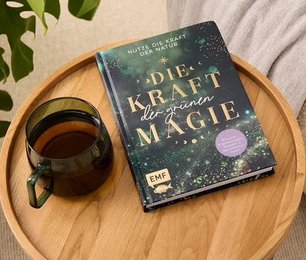 Sur un plateau en bois reposent un livre intitulé "Die Kraft der grünen Magie" et une tasse en verre contenant une boisson.
