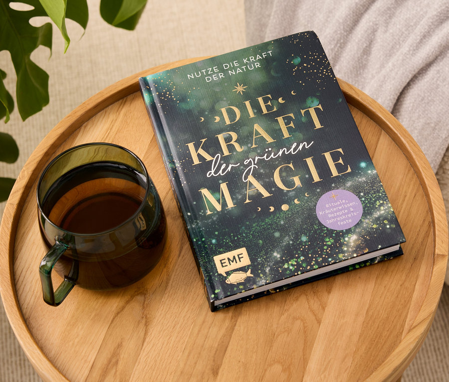 Sur un plateau en bois reposent un livre intitulé "Die Kraft der grünen Magie" et une tasse en verre contenant une boisson.