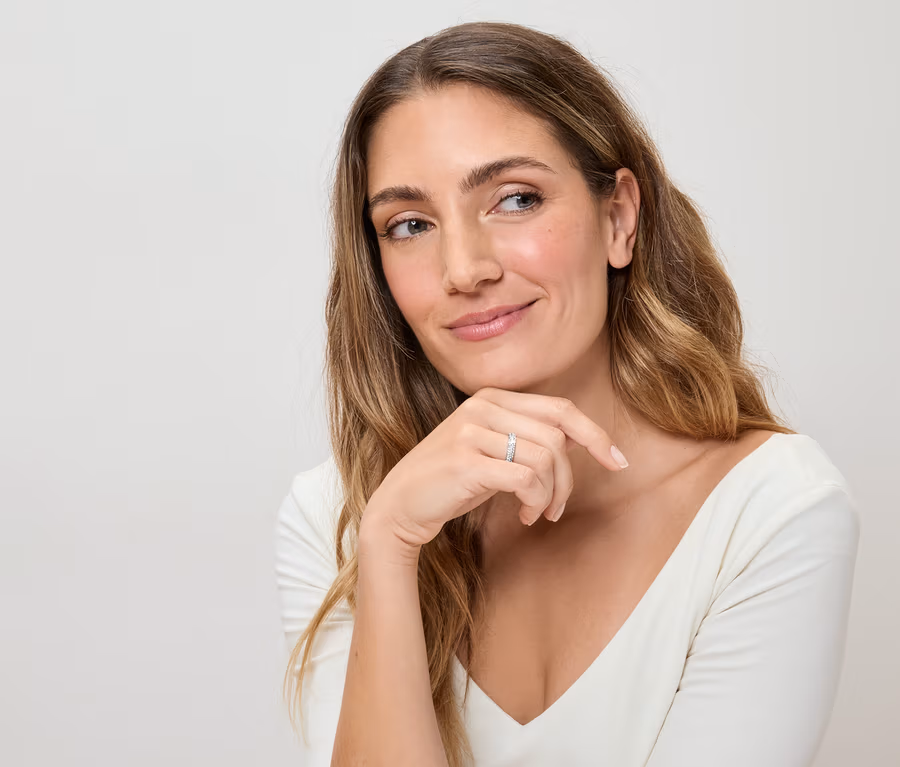 Femme aux longs cheveux ondulés et haut blanc, portant une bague argentée au doigt, regardant pensivement sur le côté.