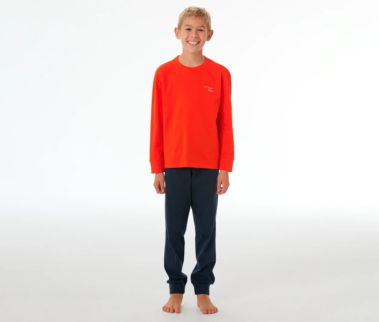 Garçon souriant portant un haut orange à manches longues et un pantalon de pyjama bleu foncé à revers, debout pieds nus.