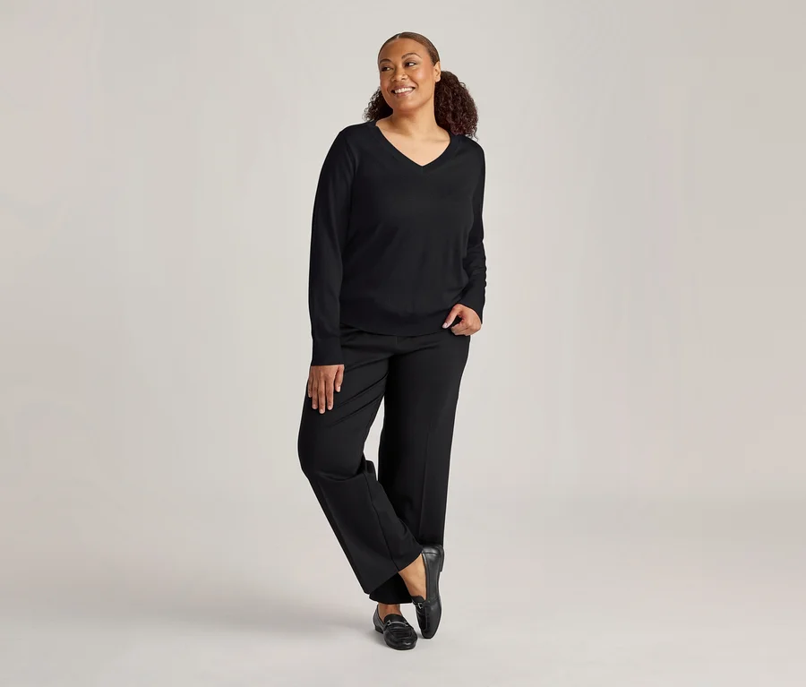 Une femme souriante aux cheveux bruns bouclés, vêtue d'un haut noir à manches longues et col en V, d'un pantalon noir et de mocassins noirs, se tient la main dans la poche et une jambe croisée.