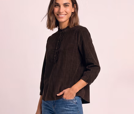 Femme souriante portant un chemisier en velours côtelé Street One marron avec col montant à volants, la main gauche dans la poche.