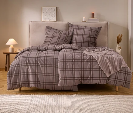 Lit double avec parure de lit en molleton à carreaux gris-brun et jeté de lit tricoté beige. À côté, une table de chevet avec lampe et bougies.