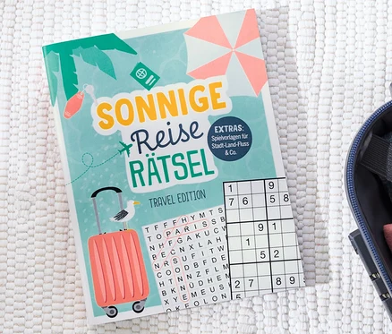 Le livre « Sonnige Reise Rätsel » est posé à côté d'une valise ouverte.