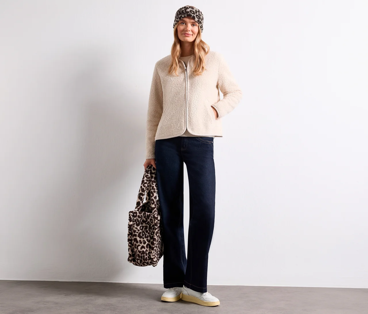 Une femme porte une veste en polaire beige, un jean foncé, des baskets blanches et un bandeau Street One à imprimé léopard, tenant un sac assorti.