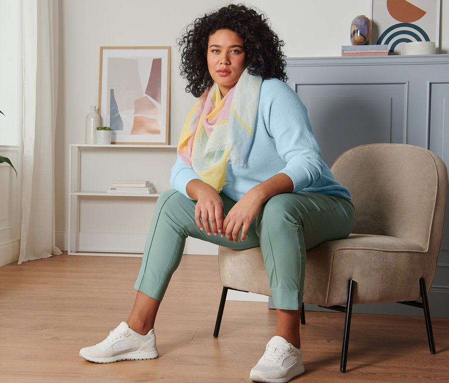 Une femme est assise sur une chaise et porte un pull en maille bleu ciel, un pantalon stretch vert menthe, une écharpe tissée et des baskets en cuir velours.