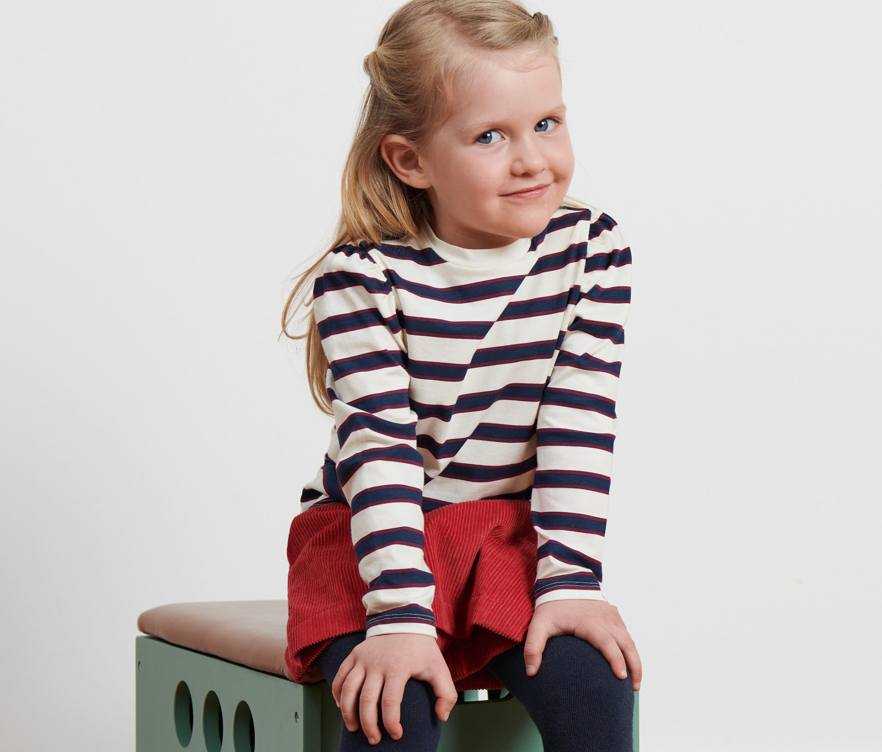 Petite fille assise sur un jouet vert, portant un T-shirt à manches longues enfant, une jupe rouge et des bottes.