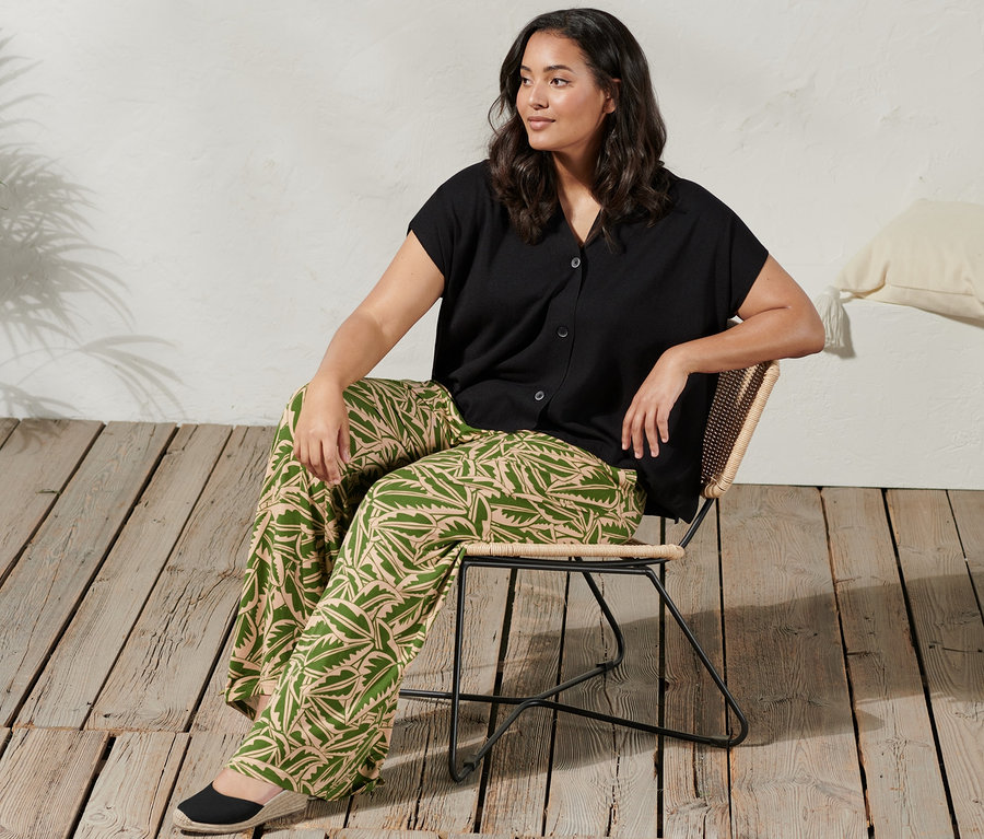 Une femme est assise sur une chaise et porte un pantalon palazzo imprimé vert beige, un cardigan à manches courtes et des espadrilles à talon compensé.