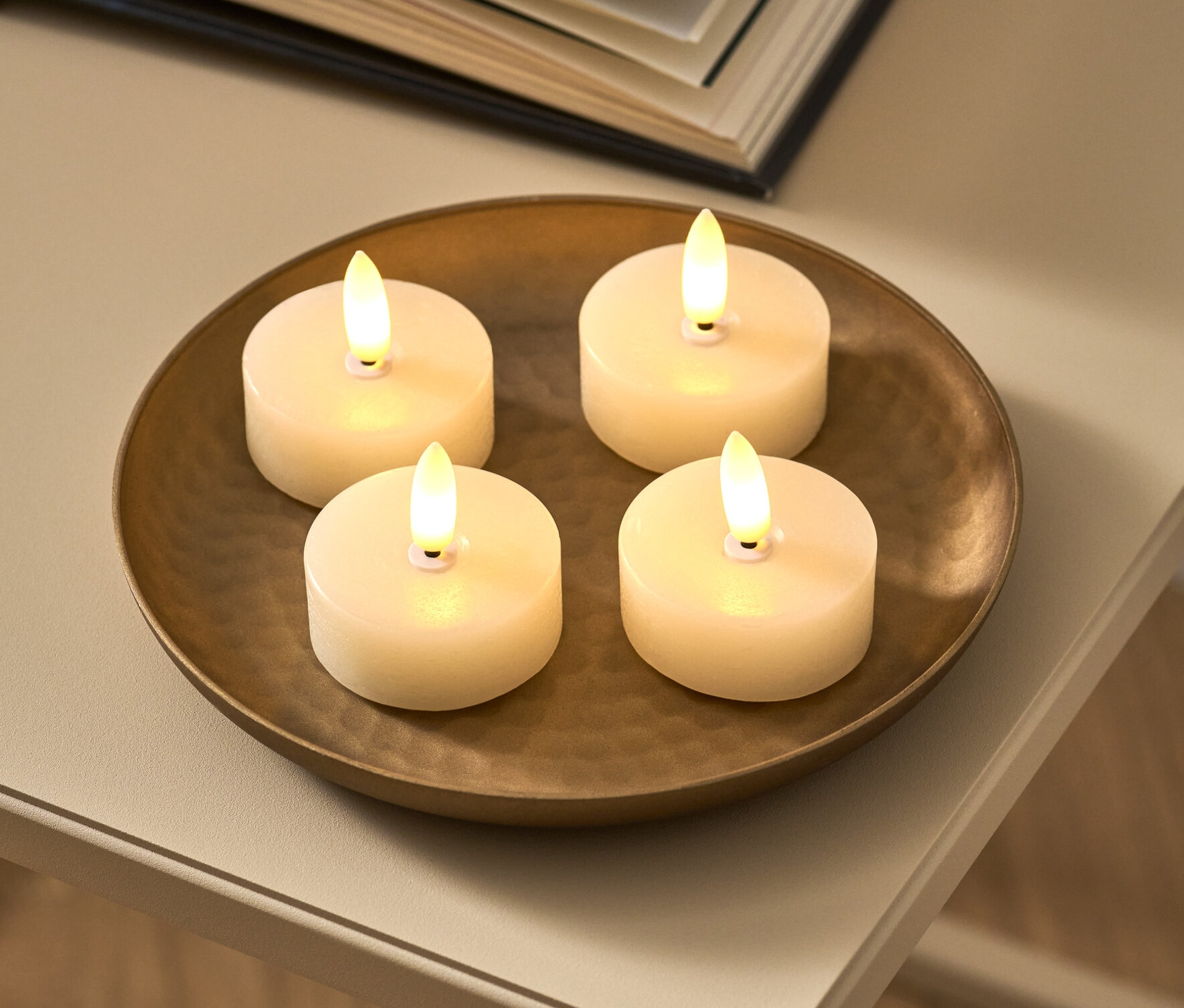 Quatre bougies chauffe-plat en cire avec LED, blanc crème, sont posées sur un plat bronze. À côté, un livre ouvert.