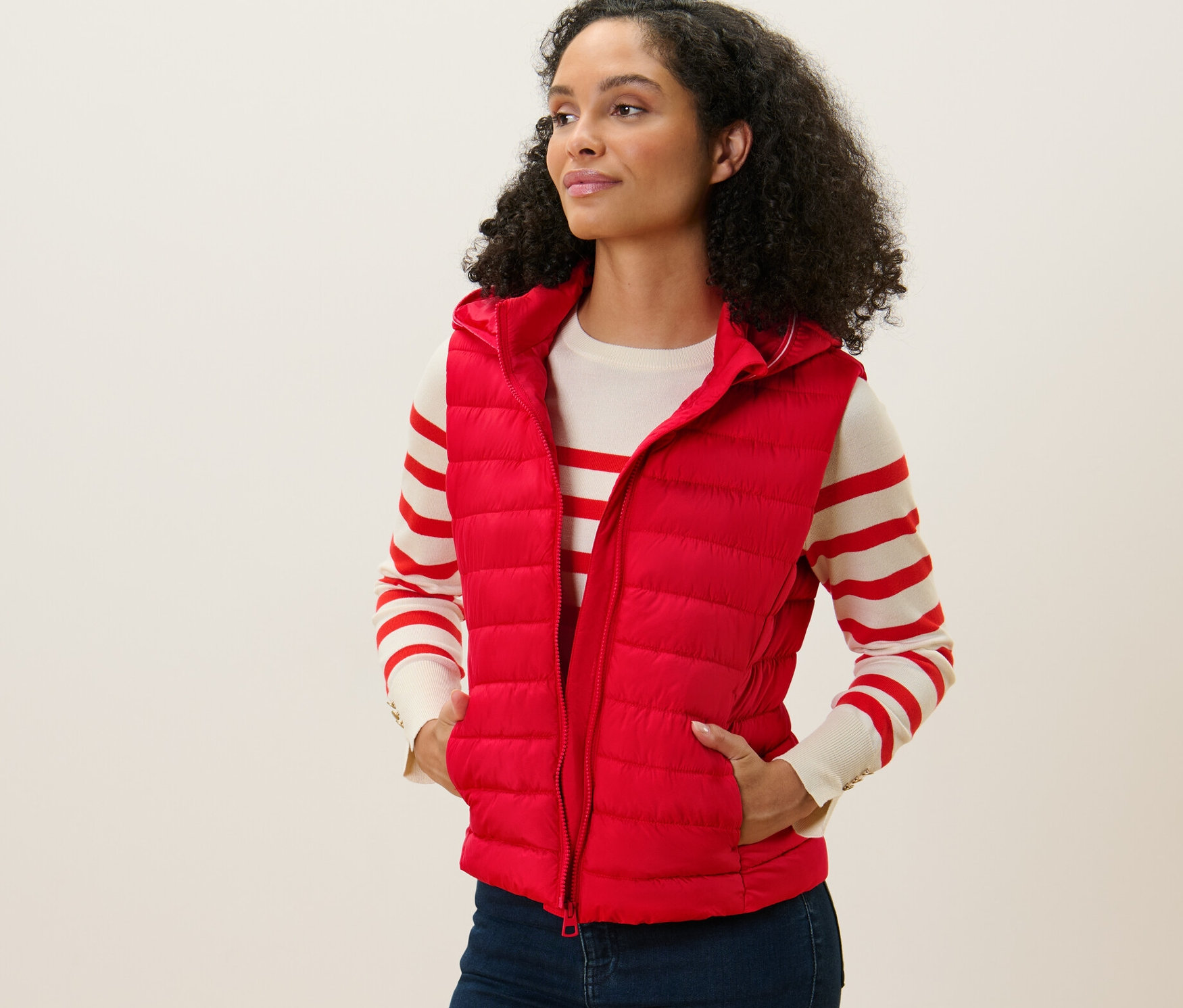 Une femme porte un gilet sans manches matelassé à capuche rouge, elle est debout et regarde vers la gauche.
