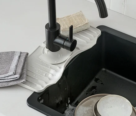 Une étagère en silicone pour robinet est placée sous un robinet noir. De la vaisselle sale se trouve dans l'évier.