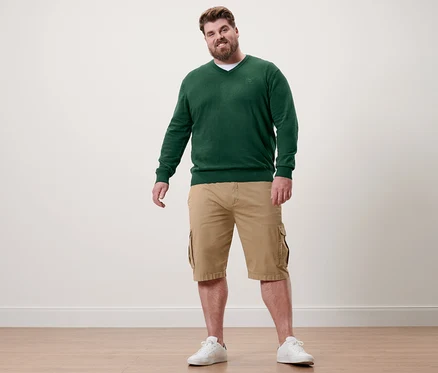 Un homme debout porte un pull vert foncé à col en V, un short kaki et des baskets blanches.