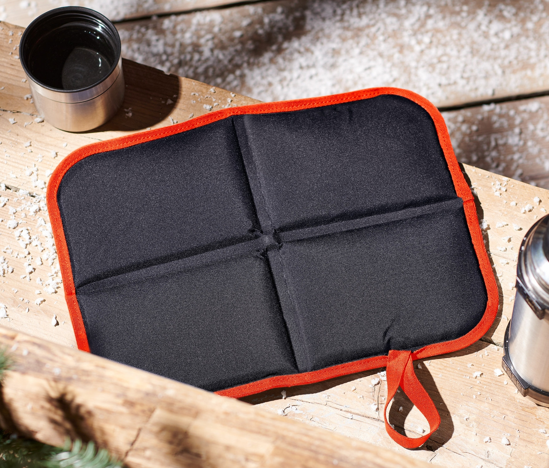 Sur une surface en bois se trouvent un coussin de siège pliable, un thermos et une tasse.