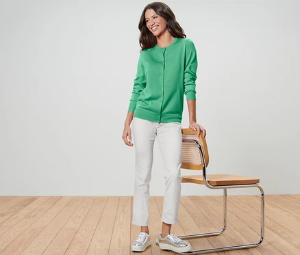 Une femme se tient appuyée sur une chaise, portant un cardigan en maille fine vert, un pantalon blanc et des chaussures argentées.