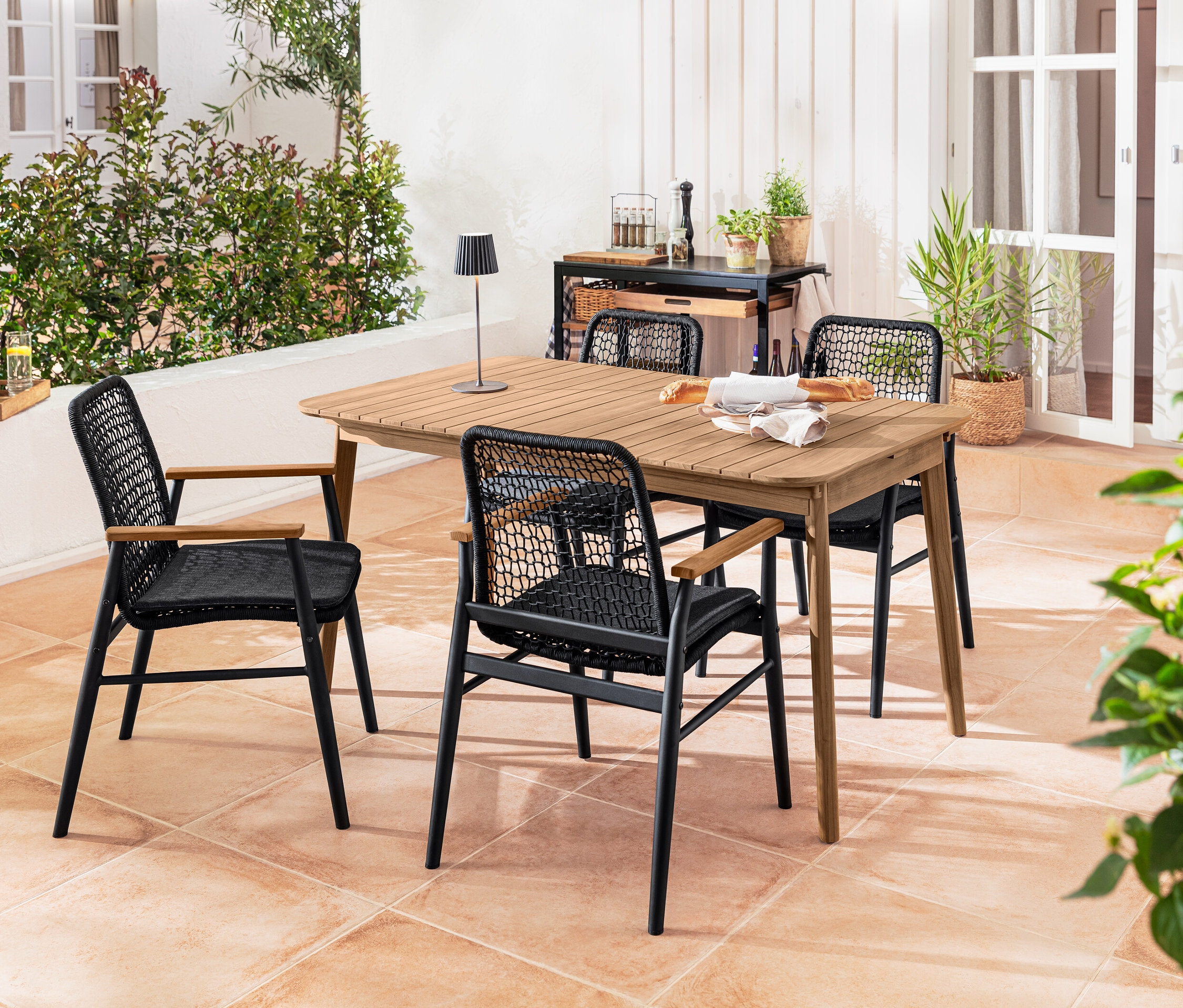 Quatre chaises Dining avec accoudoirs en teck et tressage textile sont disposées autour d'une table à rallonge en teck. Une lampe de table d’extérieur à LED rechargeable noire est posée sur la table.