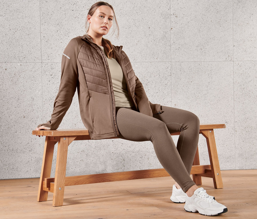 Une femme assise sur un banc porte une veste thermique à capuche marron, un t-shirt fonctionnel manches longues taupe et un legging de course à pied premium sans coutures.